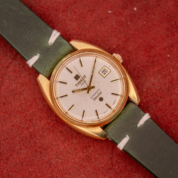 A Tissot Seastar Automatic Date Ref. 44660 Cal. 786-2 c.1970s with a green leather strap and gold case sits on a red textured surface, showcasing vintage style.