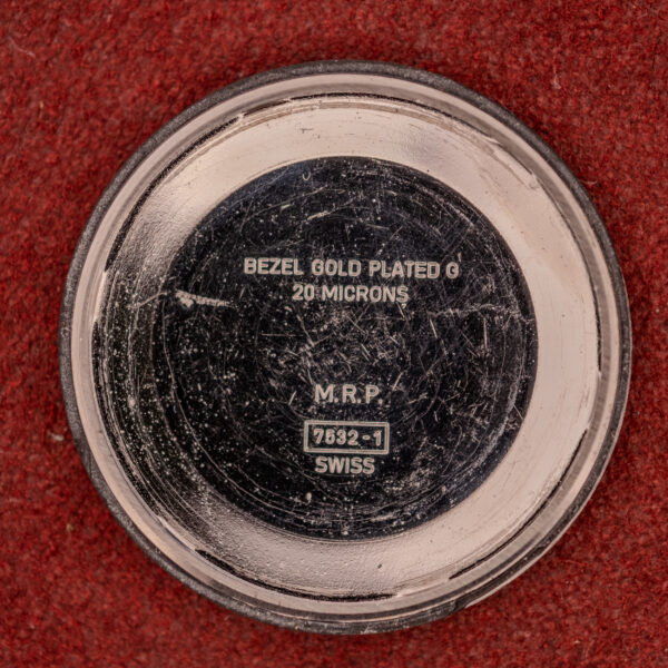 A round watch back marked BEZEL GOLD PLATED G 20 MICRONS, from a Bulova Automatic Ambassador Day-Date Ref. 7532-1 Cal. 11AOACB c.1970s, rests on a red textured surface.