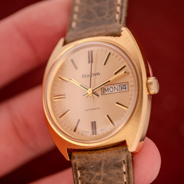 A Bulova Automatic Ambassador Day-Date Ref. 7532-1 Cal. 11AOACB from the 1970s, featuring a gold case, brown leather strap, and day-date display, is shown held in a hand.