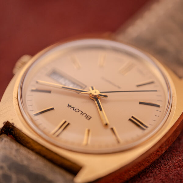 Close-up of a gold Bulova Automatic Ambassador Day-Date Ref. 7532-1 Cal. 11AOACB (c.1970s) wristwatch with leather strap, showing the time and day on a textured background.