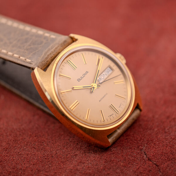 A Bulova Automatic Ambassador Day-Date Ref. 7532-1 Cal. 11AOACB c.1970s with a brown leather band is displayed elegantly on a red textured surface.