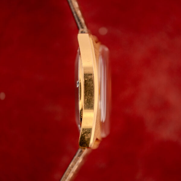 A Bulova Automatic Ambassador Day-Date Ref. 7532-1 Cal. 11AOACB c.1970s gold watch case is shown from the side against a textured red background, highlighting the refined style of this classic timepiece.