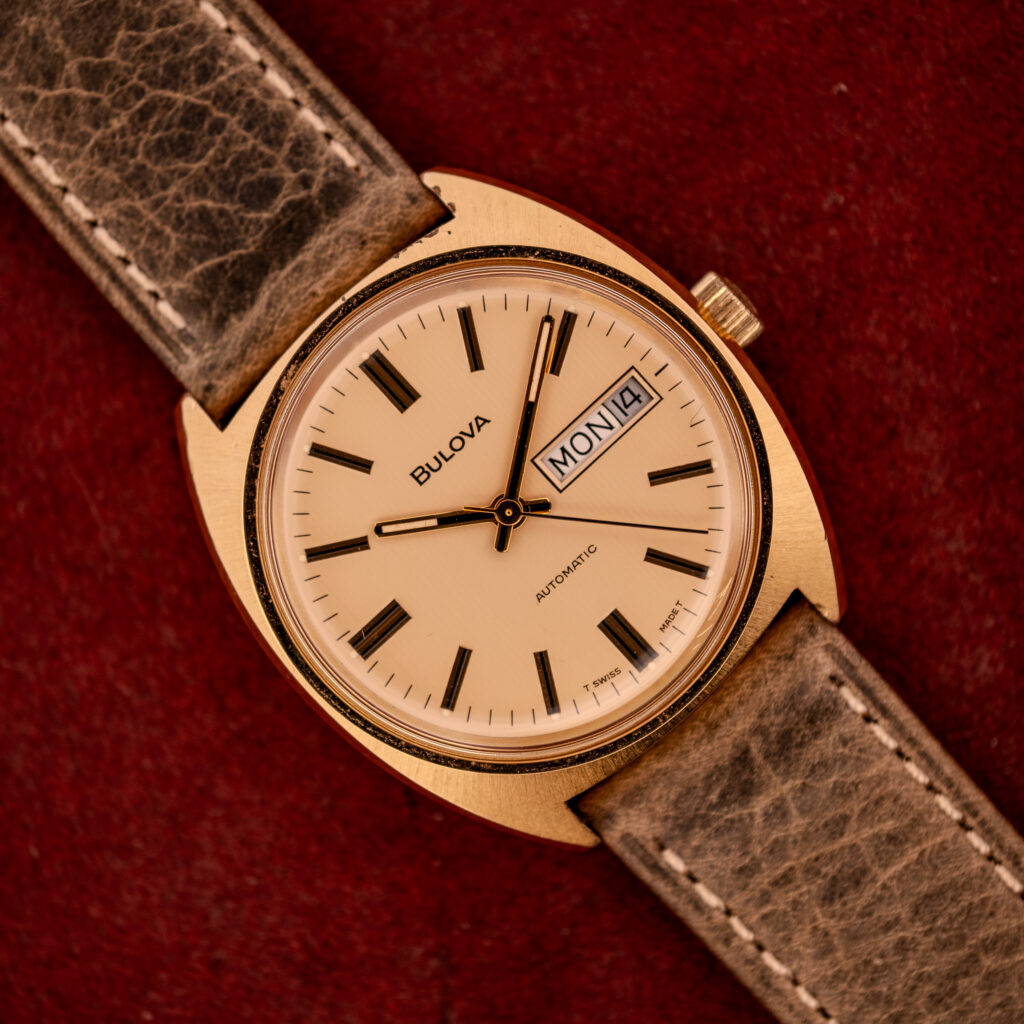 The Bulova Automatic Ambassador Day-Date Ref. 7532-1 Cal. 11AOACB, c.1970s, features a gold case, brown strap, and classic Day-Date display, set against a red textured background for vintage appeal.