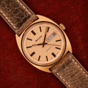 The Bulova Automatic Ambassador Day-Date Ref. 7532-1 Cal. 11AOACB, c.1970s, features a gold case, brown strap, and classic Day-Date display, set against a red textured background for vintage appeal.