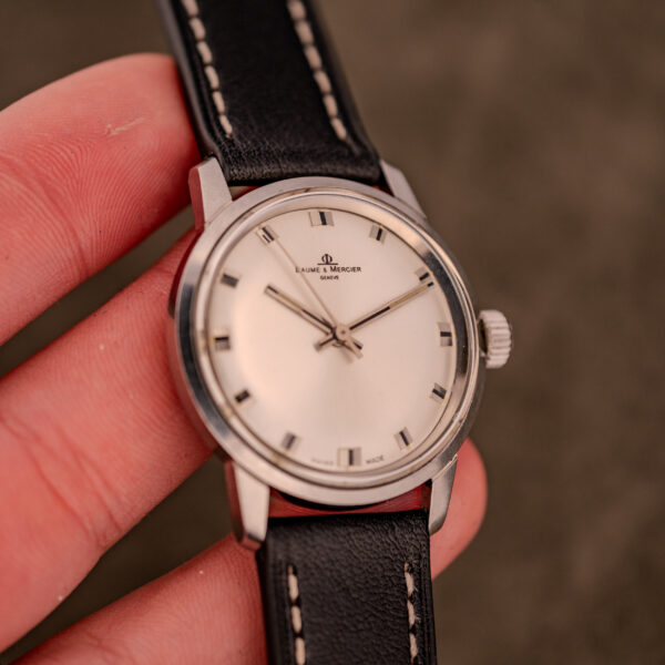 A hand holding a Baume & Mercier Compressor Manual Wind Ref. 1193 Cal. 391 c.1967 wristwatch, with a black leather strap and sleek silver dial.