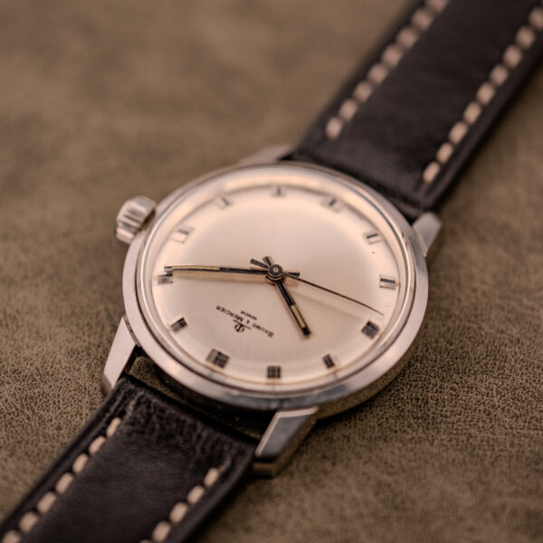 A Baume & Mercier Compressor Manual Wind Ref. 1193 Cal. 391 c.1967 wristwatch with a cream dial and black leather strap rests on a textured brown surface, showcasing its classic vintage design.