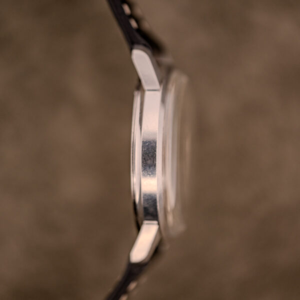 Side close-up of a Baume & Mercier Compressor Manual Wind Ref. 1193 Cal. 391 c.1967 wristwatch with a metal case and black strap, set against a blurred brown background.