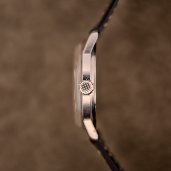 Side view of a Baume & Mercier Compressor Manual Wind Ref. 1193 Cal. 391 c.1967 silver watch case and textured crown, set against a brown blurred background.