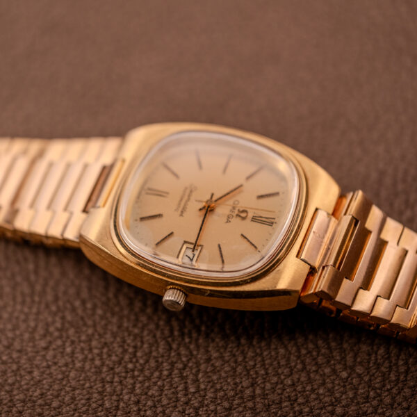 An Omega Seamaster Automatic Date "TV Case" Ref. 166.0207 Cal. 1010 gold wristwatch with a metal band is displayed on a textured brown surface.