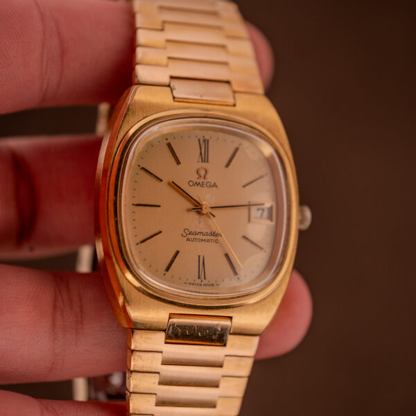 Omega Seamaster Automatic Date "TV Case" Ref. 166.0207 Cal. 1010 wristwatch held between fingers against a brown background.