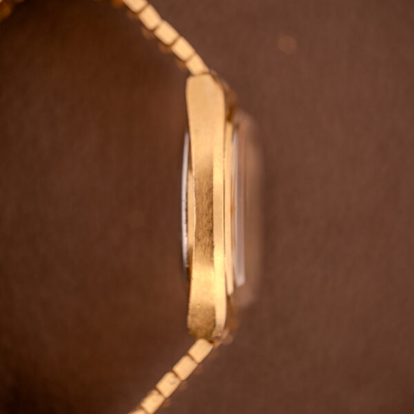 Side view of an Omega Seamaster Automatic Date "TV Case" Ref. 166.0207 Cal. 1010 gold wristwatch with a metallic band, set against a brown background.