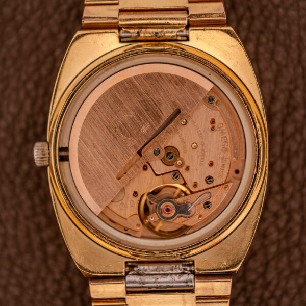A close-up of the Omega Seamaster Automatic Date "TV Case" Ref. 166.0207 Cal. 1010, gold wristwatch with exposed mechanical movement on a brown textured background.