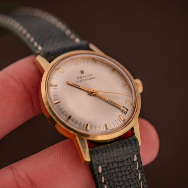Close-up of a Zenith Automatic Date Cal. 2542 watch with a sunburst dial, gold case, and black leather strap, held between fingers.