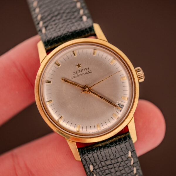 A hand holds the Zenith Automatic Date Cal. 2542, a 25-jewel wristwatch with a gold case, black leather strap, and striking silver sunburst dial.