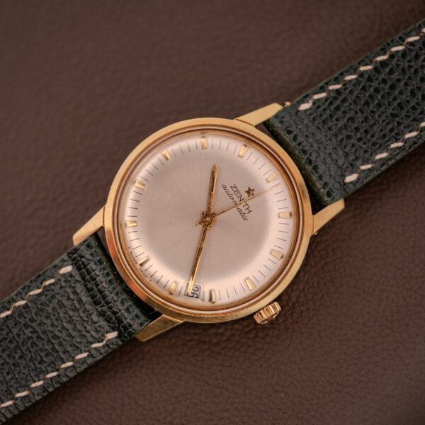 The Zenith Automatic Date Cal. 2542 25 Jewels Sunburst Dial watch features a silver sunburst dial and green leather strap, stylishly showcased on a brown textured surface.