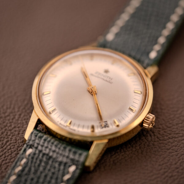 A Zenith Automatic Date Cal. 2542 25 Jewels Sunburst Dial gold wristwatch with a green leather strap is displayed on a brown textured surface.