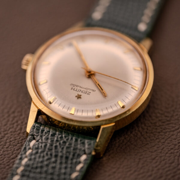 Close-up of a Zenith Automatic Date Cal. 2542 25 Jewels wristwatch with a green leather strap and sunburst dial, resting on a brown surface.