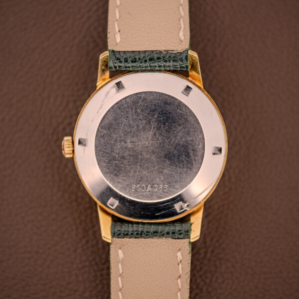 A close-up of the scratched back of a gold Zenith Automatic Date Cal. 2542 25 Jewels Sunburst Dial with a green and beige leather strap, resting on a brown surface.