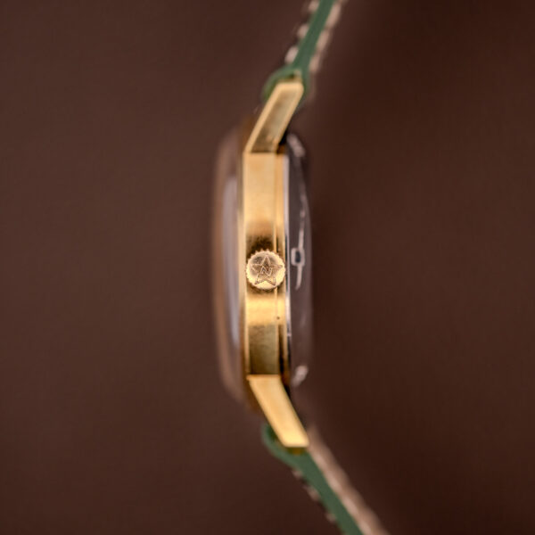 Close-up side view of a Zenith Automatic Date Cal. 2542 25 Jewels Sunburst Dial gold wristwatch with a textured crown and green leather strap against a brown background.