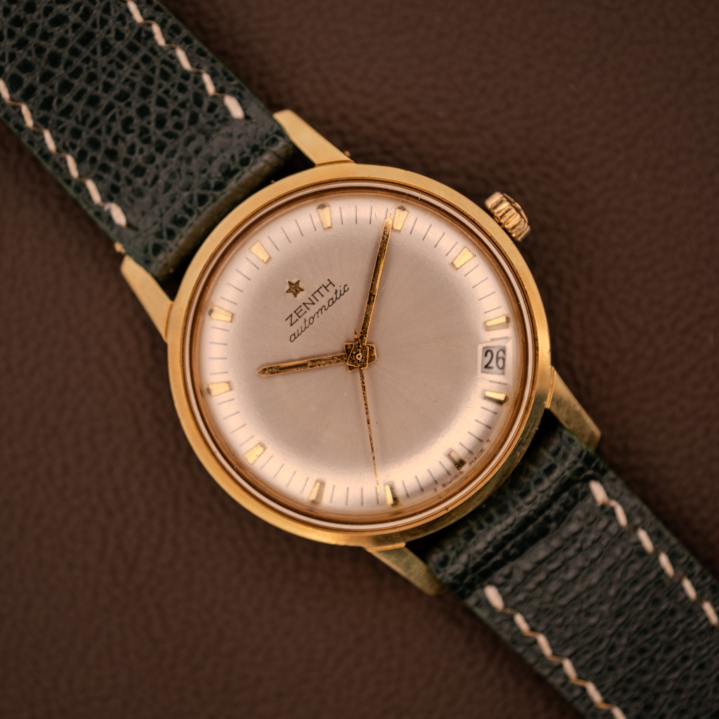 A vintage Zenith Automatic Date Cal. 2542 25 Jewels wristwatch with a sunburst dial and black leather strap, displayed on a brown surface. A vintage Zenith Automatic Date Cal. 2542 25 Jewels wristwatch with a sunburst dial and black leather strap, displayed on a brown surface.