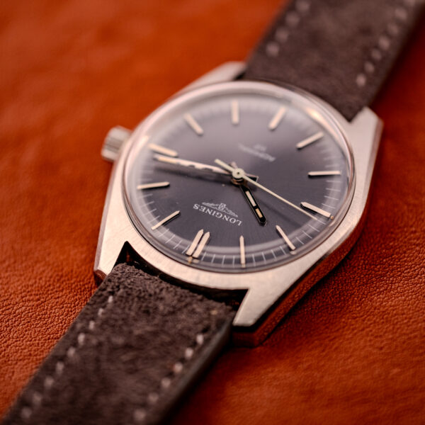 A Longines Admiral HF Anthracite Sunburst Dial Ref. 2301 Cal. 6942 c.1972 with a suede strap rests elegantly on a brown leather surface.