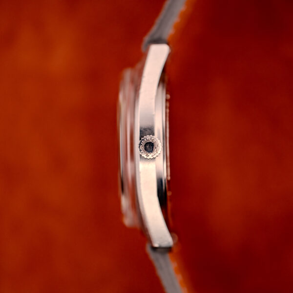 Side view of a Longines Admiral HF Anthracite Sunburst Dial Ref. 2301, Cal. 6942 c.1972, silver case with crown, set against a blurred orange background; hints of the signature sunburst dial are visible.