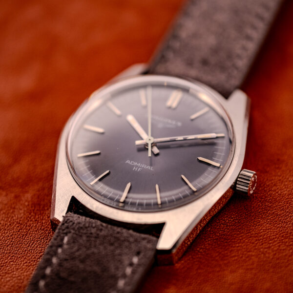 Close-up of a Longines Admiral HF Anthracite Sunburst Dial Ref. 2301 Cal. 6942 c.1972 wristwatch with silver case and suede gray strap, shown on a brown surface.