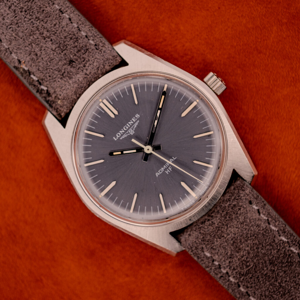 A Longines Admiral HF Ref. 2301 (c.1972) with an anthracite sunburst dial and suede strap, Cal. 6942, displayed against a vibrant orange background.
