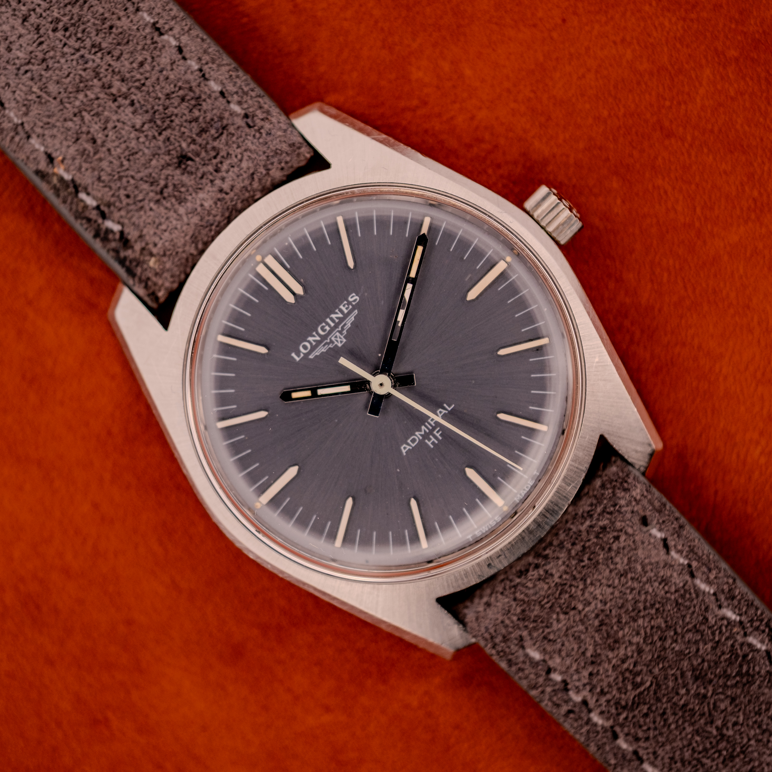 A Longines Admiral HF Ref. 2301 (c.1972) with an anthracite sunburst dial and suede strap, Cal. 6942, displayed against a vibrant orange background. A Longines Admiral HF Ref. 2301 (c.1972) with an anthracite sunburst dial and suede strap, Cal. 6942, displayed against a vibrant orange background.