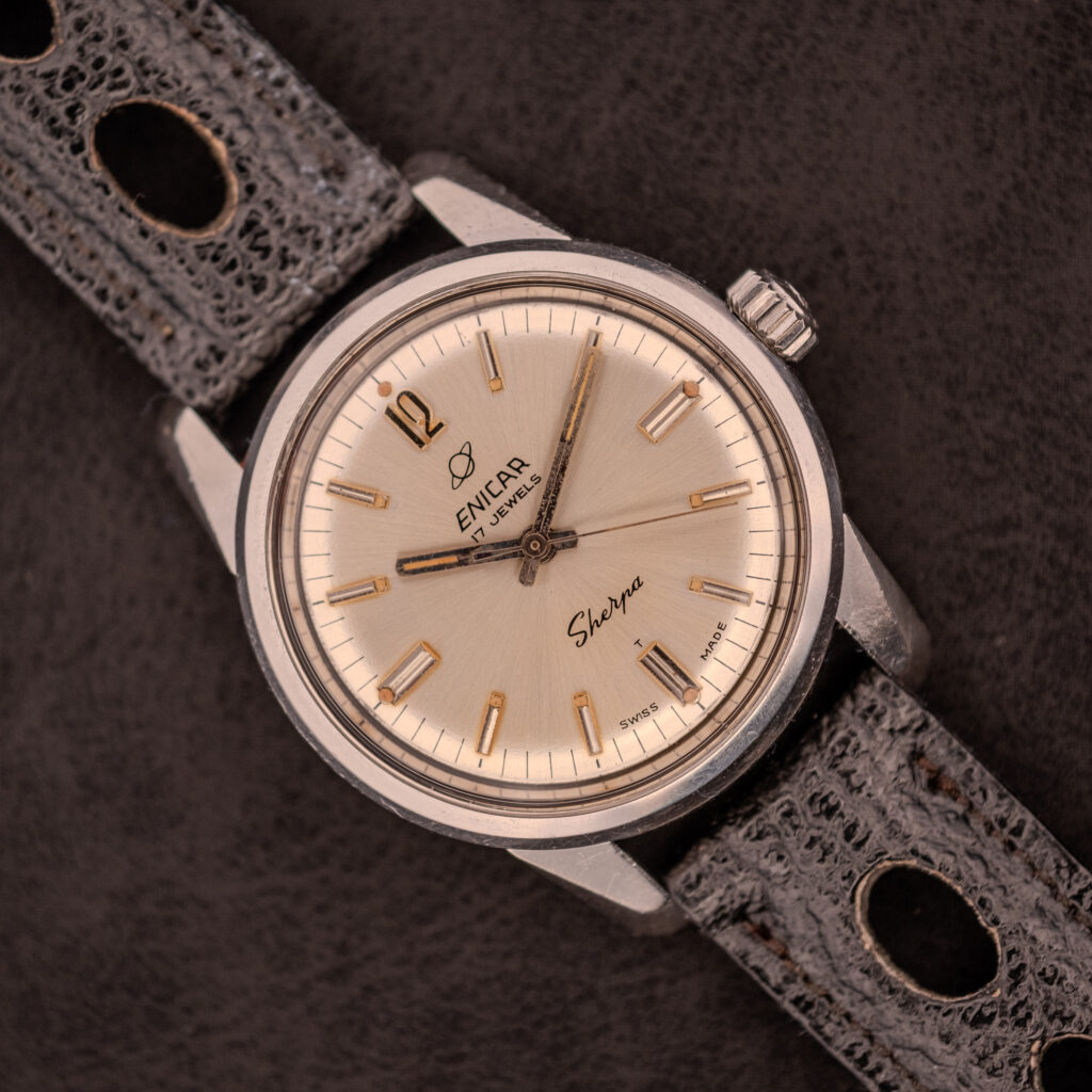 An Enicar Sherpa Seapearl 600 Super Compressor Ref. 140/013 Cal. AR 1140, featuring a beige dial and black perforated leather strap, highlights classic Super Compressor design on a dark surface.