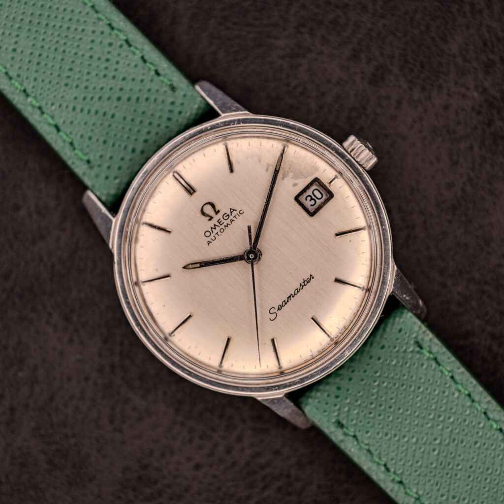 A vintage Omega Seamaster Automatic Date Ref. 166.037 c.1968 featuring a Cal. 565 movement, silver dial, and green strap, displayed on a black background.
