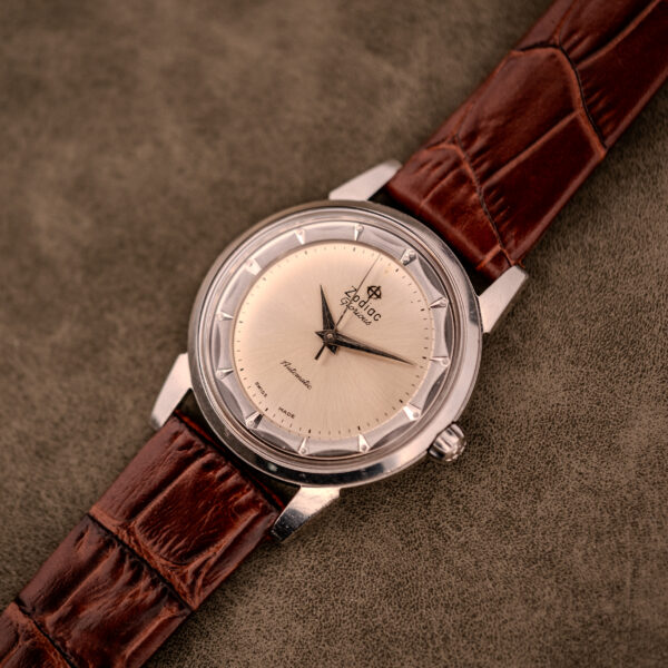 A Zodiac Glorious Automatic "Faceted Bezel" Ref. 696 Cal. 1624 silver wristwatch with a brown leather strap is displayed on a textured beige surface.