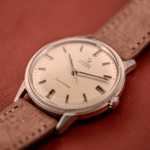 An Omega Seamaster Automatic "Red Crosshair" Ref. 165.002 Cal. 552 with a brown leather strap rests on a reddish-brown surface, showcasing its distinctive red crosshair detailing.