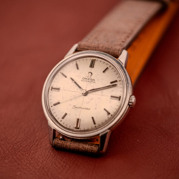 An Omega Seamaster Automatic "Red Crosshair" Ref. 165.002 Cal. 552 with a brown leather strap rests on a reddish-brown surface, featuring classic Red Crosshair accents on its dial.