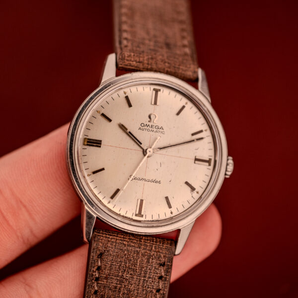 A hand holds the Omega Seamaster Automatic "Red Crosshair" Ref. 165.002 Cal. 552, featuring a brown strap and vibrant red crosshair dial, set against a vivid red background.