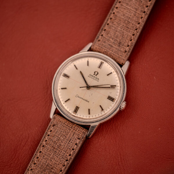 An Omega Seamaster Automatic "Red Crosshair" Ref. 165.002 Cal. 552 with a brown leather strap is displayed elegantly on a red leather surface.