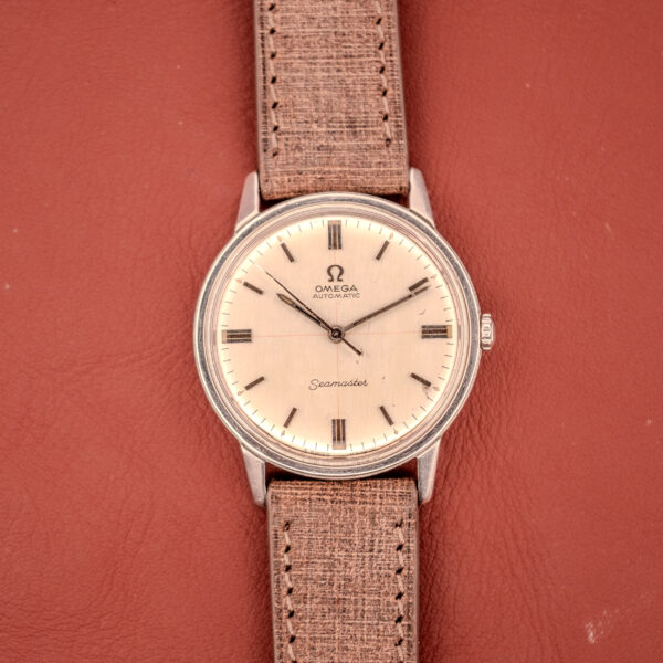 An Omega Seamaster Automatic "Red Crosshair" Ref. 165.002 Cal. 552 with a tan strap rests on a reddish-brown leather surface, showcasing its distinctive red crosshair detail.