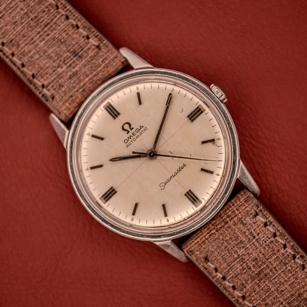 An Omega Seamaster Automatic "Red Crosshair" Ref. 165.002 Cal. 552 with a red crosshair dial and tan textured strap, displayed on reddish-brown leather.