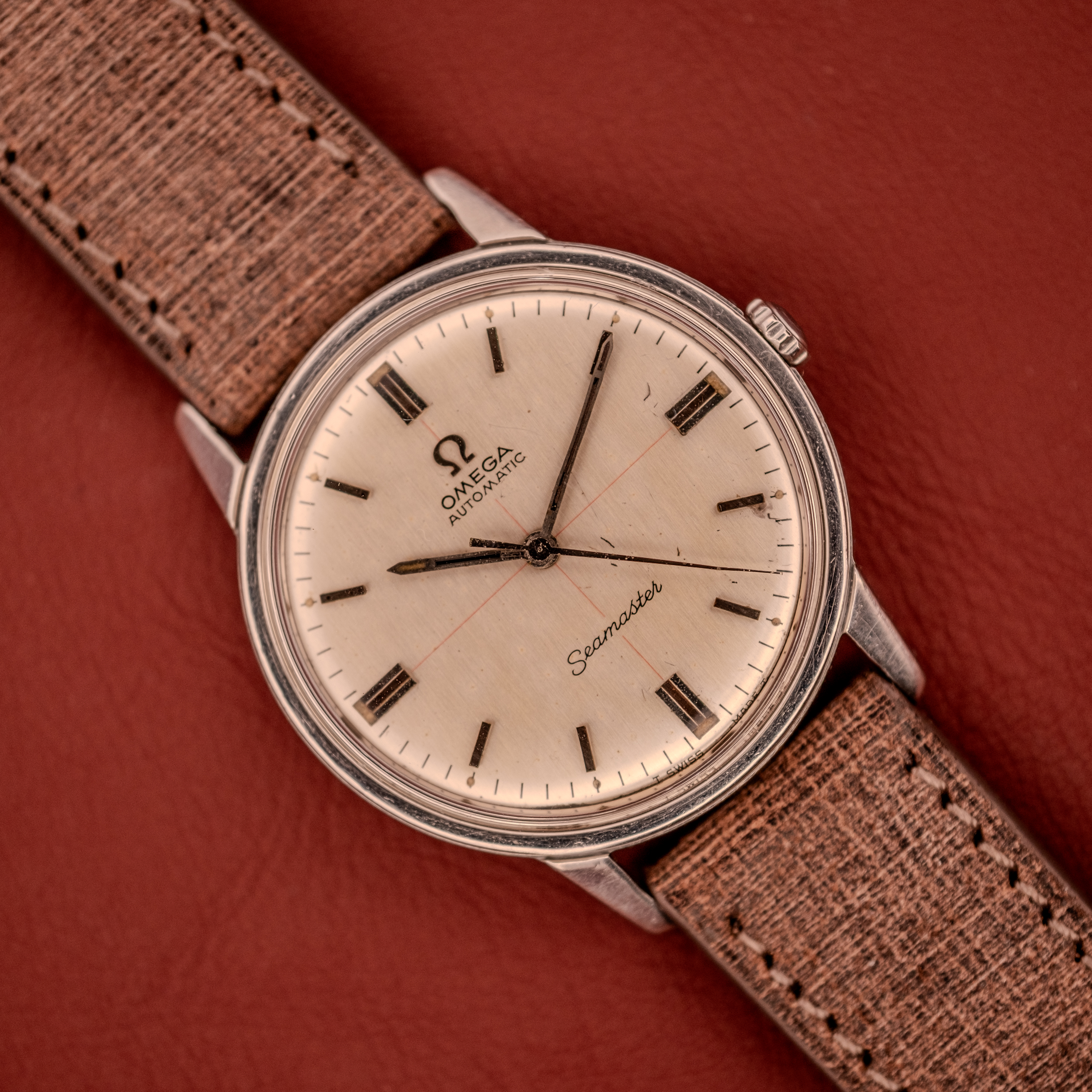 An Omega Seamaster Automatic "Red Crosshair" Ref. 165.002 Cal. 552 with a red crosshair dial and tan textured strap, displayed on reddish-brown leather. An Omega Seamaster Automatic "Red Crosshair" Ref. 165.002 Cal. 552 with a red crosshair dial and tan textured strap, displayed on reddish-brown leather.