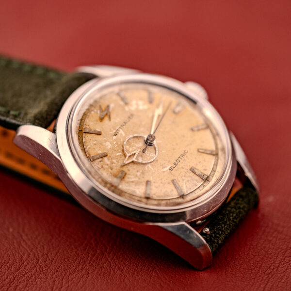 Close-up of a vintage Wittnauer Electro-Chron "Lightning Bolt" Tropic Dial Ref. 6150 with a worn beige dial and dark green strap, displayed on a red surface.