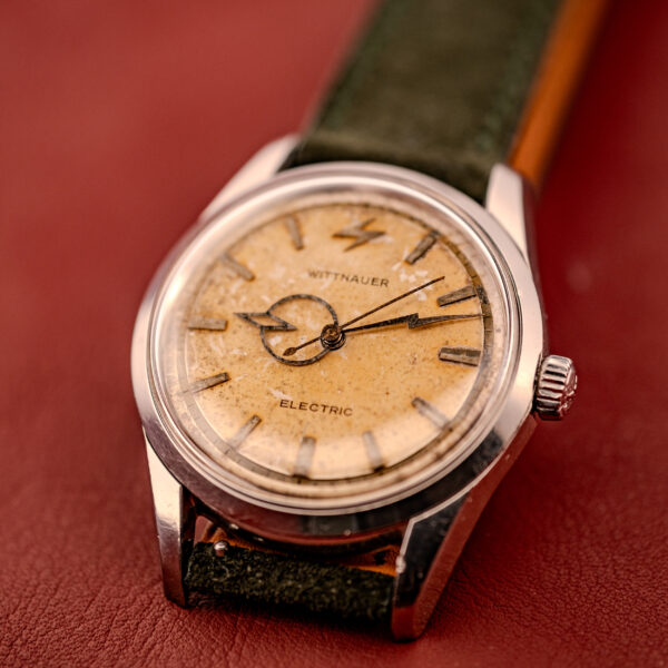 Close-up of a vintage Wittnauer Electro-Chron "Lightning Bolt" Tropic Dial Ref. 6150 wristwatch with a worn tropic dial and green leather strap, displayed on a red surface.