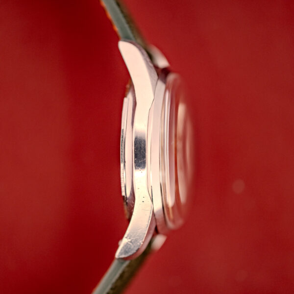 Side view of a Wittnauer Electro-Chron "Lightning Bolt" Tropic Dial Ref. 6150 wristwatch with a metal case and green strap, set against a red background.