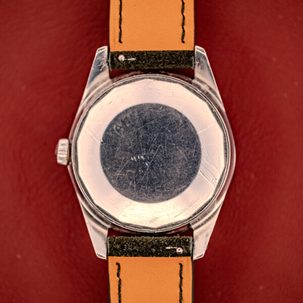 A close-up of the scratched back of a Wittnauer Electro-Chron "Lightning Bolt" Tropic Dial Ref. 6150 wristwatch with a tan leather strap on a red background.