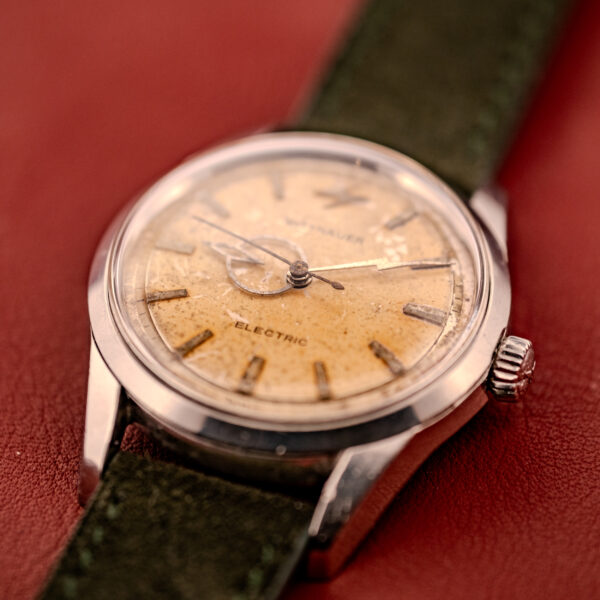 A Wittnauer Electro-Chron "Lightning Bolt" Tropic Dial Ref. 6150 wristwatch with a worn dial and green strap, displayed on a red surface.