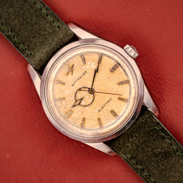 A Wittnauer Electro-Chron "Lightning Bolt" Tropic Dial Ref. 6150 wristwatch with a tan dial and green leather strap displayed on a red surface.