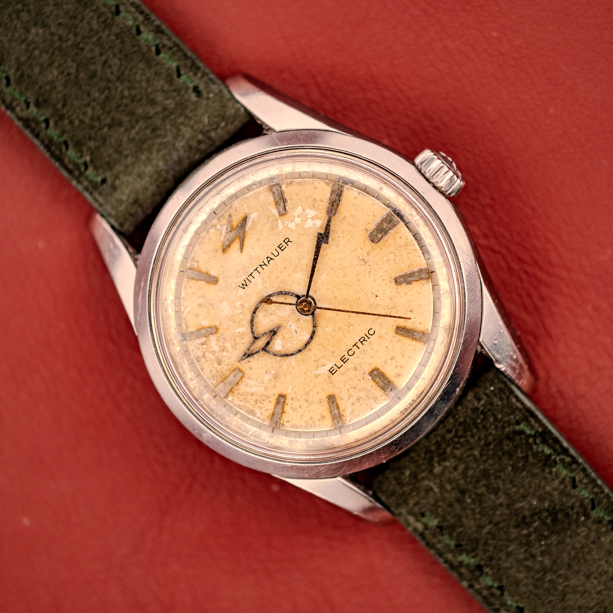 A Wittnauer Electro-Chron "Lightning Bolt" Tropic Dial Ref. 6150 wristwatch with a tan dial and green leather strap displayed on a red surface. A Wittnauer Electro-Chron "Lightning Bolt" Tropic Dial Ref. 6150 wristwatch with a tan dial and green leather strap displayed on a red surface.