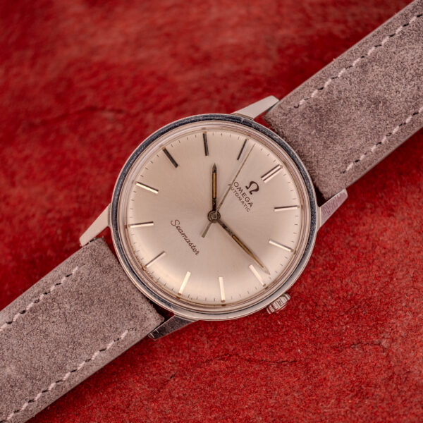 An Omega Seamaster Automatic Ref. 165.002 with Cal. 552 movement and a grey suede strap, displayed on a textured red background.