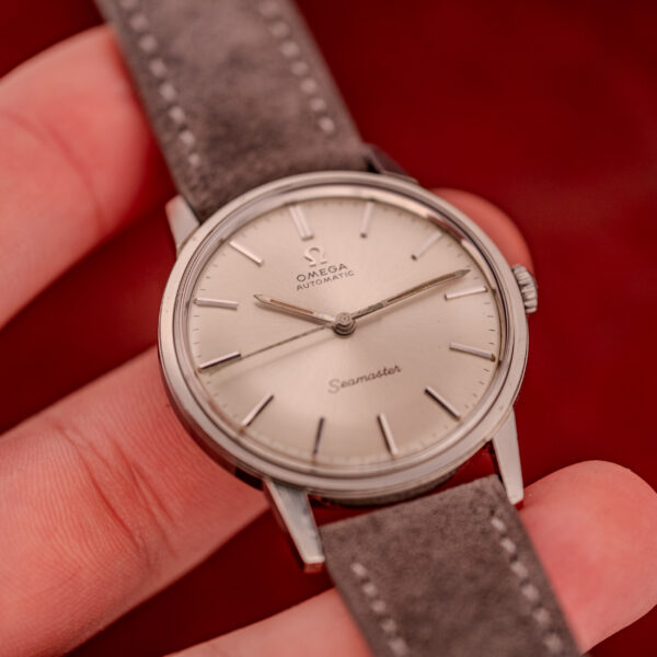 A hand holds an Omega Seamaster Automatic Ref. 165.002 Cal. 552 wristwatch with a gray leather strap against a red background.