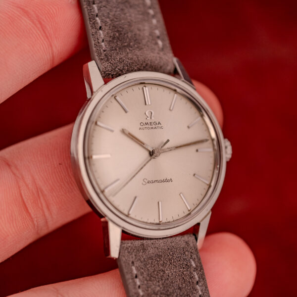 A hand holds an Omega Seamaster Automatic Ref. 165.002 Cal. 552 with a grey suede strap and silver dial, set against a red background.