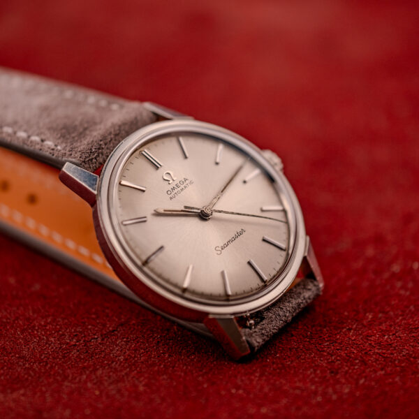 The Omega Seamaster Automatic Ref. 165.002 Cal. 552 wristwatch features a silver case, brown leather strap, and is shown on a textured red surface.
